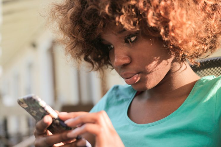  Acessibilidade Digital e Marketing inclusivo: Mulher negra com abelos cursos cacheados até o final da orelha, olhando o celular nas suas mão. Ela está de camiseta azul claro.