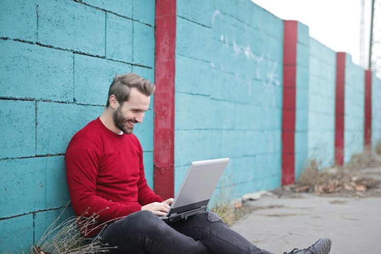 Acessibilidade Digital e Marketing inclusivo: Homem branco com barba castanha e cabelos curtos castanho está sentado no chão, encostado a um muro colorido azul e vermelho. Ele sorri para o notebook ao seu colo. Ele veste um suéter vermelho.