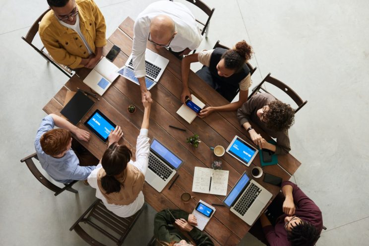 Espaços Corporativos e Tecnologia: 8 pessoas em uma reunião de negócios em volta de uma mesa de madeira, com diversos dispositivos sobre a mesa. Duas pessoas estão apertando as mãos.